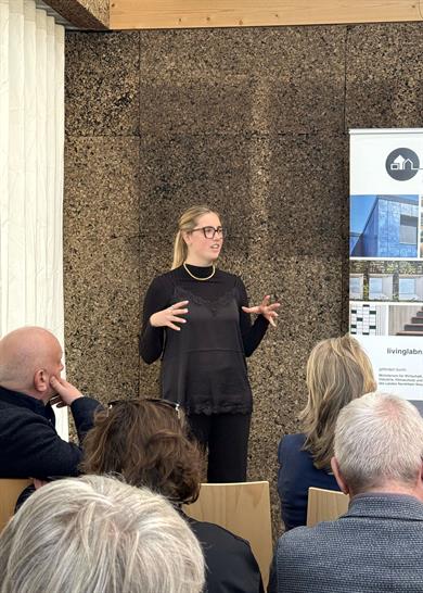 Eine Frau spricht vor sitzendem Publikum in einem Raum mit Korkwand; rechts steht ein Roll-up mit Architekturabbildungen.